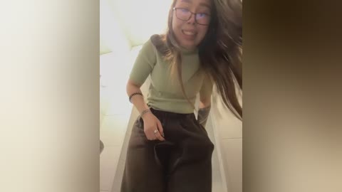 Media: A video of a smiling young woman with glasses, long brown hair, and a light green top, standing in a dimly lit room. She wears black leather pants and a beige coat, with a white wall and a door in the background.