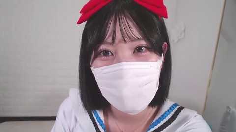 Media: Video of a young Asian woman with short black hair, wearing a white mask and sailor dress with a red bow, standing indoors against a white background.