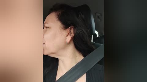 Media: Video of a middle-aged woman with dark hair, wearing a black top, seated in a car with a seatbelt on, looking distressed, eyes closed, in a dimly lit room.