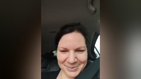 Media: Video of a smiling woman with fair skin, dark hair pulled back, wearing a black top, seated in a car with a gray seat.