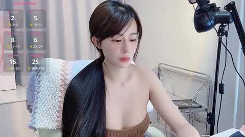 Media: A video shows a fair-skinned, young East Asian woman with long, straight black hair, wearing a strapless brown dress, sitting in front of a white chair. A large camera is on a stand to the right.