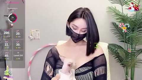 Media: Video of a light-skinned East Asian woman with straight black hair, wearing a black face mask and sheer black top, holding a small white poodle. Background includes a digital thermometer, a green plant, and a colorful flower arrangement.