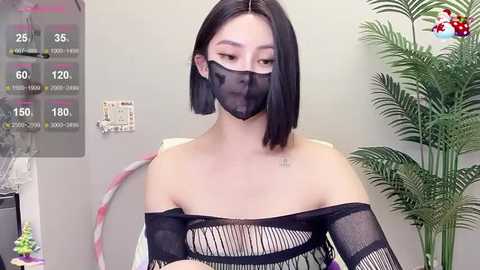 Media: A video of an East Asian woman with straight black hair, wearing a black mesh mask, off-shoulder black mesh top, and a white bra, standing in an office with a calendar and potted plant.