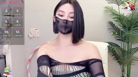 Media: Video of an Asian woman with shoulder-length black hair, wearing a black fishnet top and face mask, indoors with a calendar, potted plant, and light gray walls.