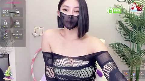 Media: Video of an Asian woman with shoulder-length black hair, wearing a black mesh top and face mask, standing indoors with a green plant in the background.