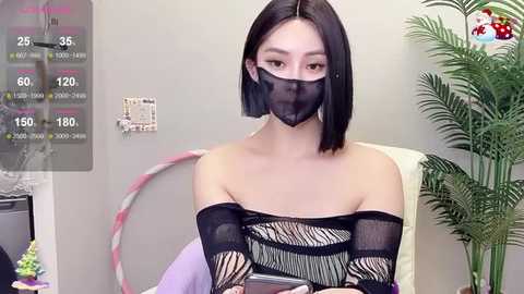 Media: Video of an East Asian woman with a black bob haircut, wearing a black mask and a black, off-shoulder, striped top, sitting at a desk in an indoor setting with a plant and a pink and white hoop.