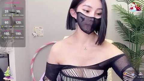 Media: Video of an East Asian woman with a short black bob haircut, wearing a black fishnet top, black mask, and pink hair ties, standing indoors with a green plant and a colorful wall background.