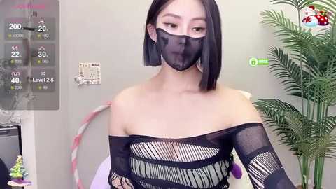 Media: Video of an East Asian woman with a pale complexion, short black bob haircut, wearing a black mesh mask and fishnet top, standing indoors near a green plant.