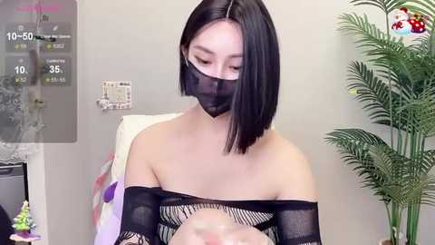 Media: Video of a young East Asian woman with a bob haircut, wearing a black mask and sheer black top, seated indoors.