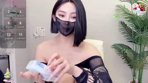 Media: Video of an East Asian woman with straight black hair, wearing a black lace mask, black lace top, and holding a blue ice pack to her left breast. Background includes a calendar, potted plant, and a light-colored wall.