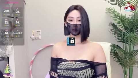 Media: Video of an Asian woman with a straight bob haircut, wearing a black mask and a black off-the-shoulder mesh top, sitting in a sterile room with a digital display and a plant.