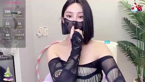 Media: A video of an East Asian woman with straight black hair, wearing a black fishnet top and a black face mask, holding a finger to her lips. The background features a wall clock, a thermometer, and a green plant.