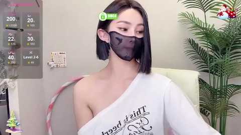 Media: Video of a young Asian woman with straight black hair, wearing a white off-shoulder shirt, black face mask, and green hair clip, seated in a medical setting with a plant and digital display screen in the background.