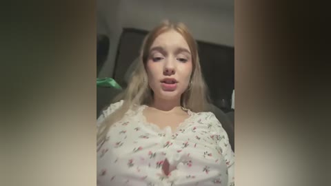 Media: Video of a young Caucasian woman with long blonde hair, wearing a floral-patterned blouse, and a nose ring. She's indoors, with a blurred background.