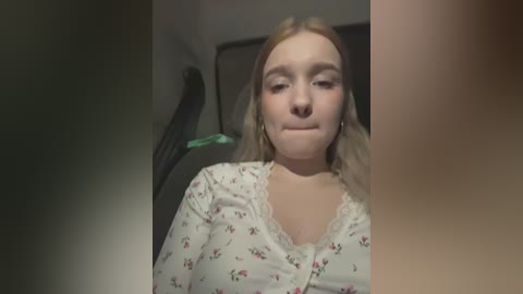 Media: Video of a fair-skinned woman with shoulder-length blonde hair, wearing a floral-patterned shirt, sitting in a dark car. She has a neutral expression, and the background is blurred.