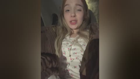 Media: A video of a young Caucasian girl with long blonde hair, wearing a floral top and a brown jacket, sitting in a dimly lit room, eyes closed, mouth slightly open.