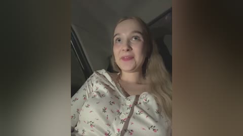 Media: Video of a young Caucasian woman with blonde hair and light skin, wearing a floral blouse, seated in a dimly lit car interior.