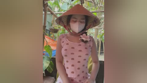 Media: Video of a young Asian woman wearing a pink polka-dotted shirt, traditional conical hat, and mask, standing in a lush garden with green foliage and bamboo structures in the background.