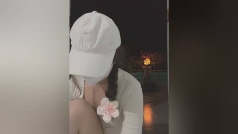 Media: A video of a woman in a white baseball cap, long braids, and a white shirt, standing in a dark room. She has a pink flower pinned to her chest. In the background, a lit fire pit glows, with blurred trees and a dark sky.