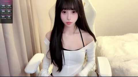 Media: Video of a young East Asian woman with long black hair, fair skin, and an off-shoulder white top, seated on a plush white gaming chair, in a minimalist room with beige curtains.