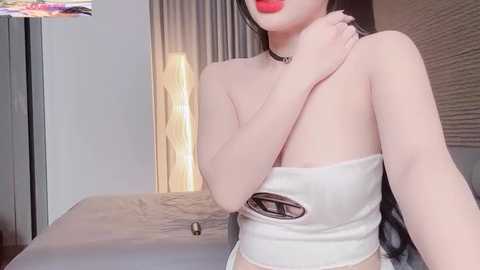 A video of an Asian woman with fair skin, red lipstick, and long black hair, wearing a white, strapless crop top and a choker, sitting on a bed in a modern bedroom with beige curtains and a lamp.