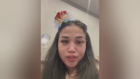 Media: Video of a young Latina woman with long dark hair, wearing a red and gold glittery tiara, makeup, and a white top.