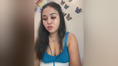 Media: Video of a young Asian woman with long black hair, wearing a blue knitted top and a glittery crown, blowing a kiss against a beige wall adorned with butterfly stickers.