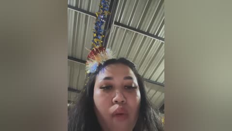 Media: Video of a young woman with long black hair, wearing a festive, colorful feathered headband and blowing a kiss, in a dimly lit, corrugated metal-roofed room.