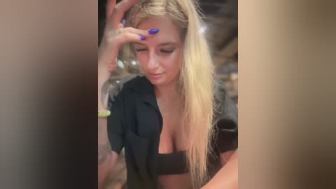 Media: Video of a blonde woman with fair skin, long hair, wearing a black shirt and black bra, touching her face, with a blurred background.