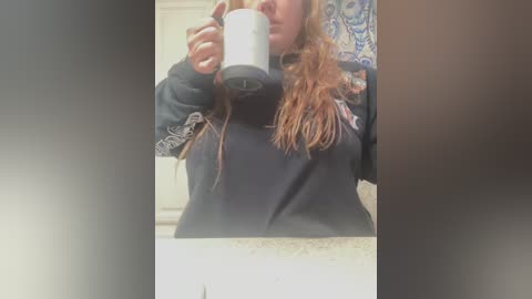 Media: A video of a woman with long, reddish-brown hair, wearing a black hoodie and holding a white mug, partially obscured by a blurred background.