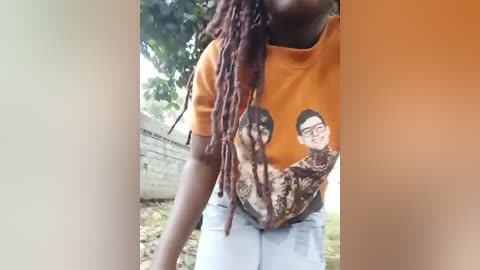 Media: Video of a young Black woman with long, brown dreadlocks, wearing an orange t-shirt with a cartoon design and light blue jeans. Background features a white brick wall and green foliage.