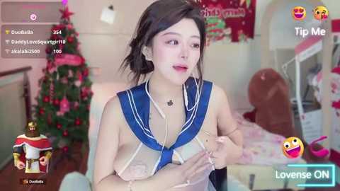 Media: A video of an Asian woman with fair skin, dark hair, and a blue sailor school uniform, holding a phone. She's in a cozy bedroom with a Christmas tree and heart-shaped decorations.