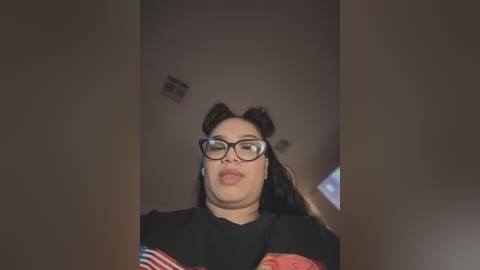 Media: Video of a young woman with fair skin, wearing black glasses and a black shirt with an American flag pattern, sticking out her tongue. She has dark hair styled in two buns.