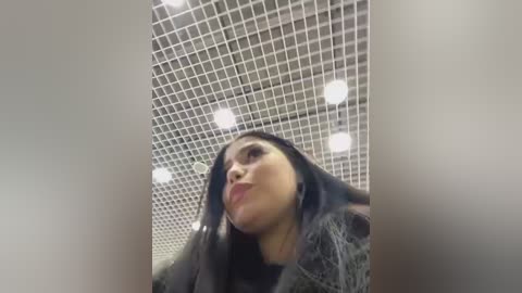 Media: Video of a woman with long, dark hair, wearing a fur coat, looking upwards at a tiled ceiling with bright lights. She has a slightly tilted head and pursed lips.