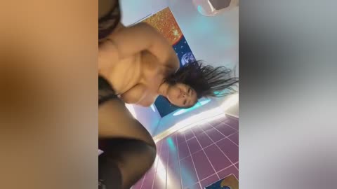 Media: A video of a topless woman with long black hair, wearing black stockings, standing in a room with pink-tiled floor and colorful wall art.