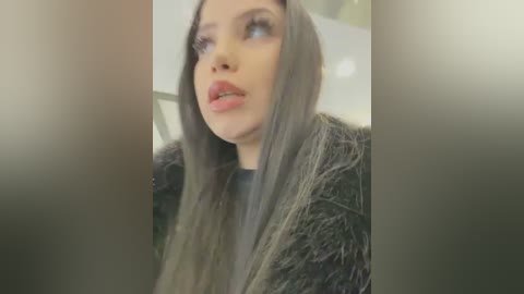 Media: A video of a young woman with long, straight, gray hair, fair skin, and light makeup, wearing a black fur coat, looking introspective. The background is blurred.