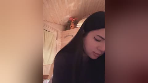 Media: Video of a young woman with long black hair, wearing a black hijab, standing in a cozy, wooden-paneled room with a wooden ceiling and a decorative shelf holding small figurines and a soft toy.