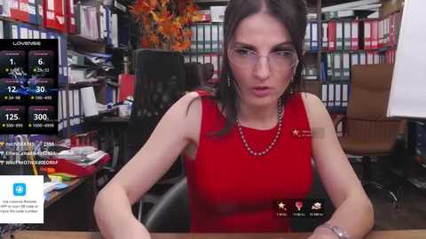 Media: Video of a woman with glasses, red dress, and necklace, sitting at a desk in a cluttered office with bookshelves and a TV displaying news and social media.