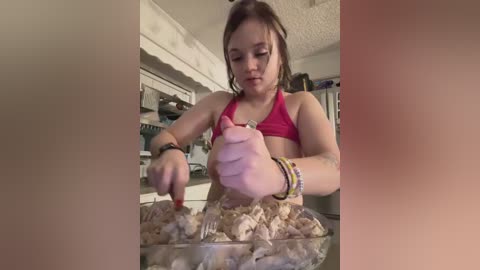 Media: Video of a young girl in a pink bikini, stirring a bowl of uncooked meatballs in a kitchen with beige walls and cabinets.