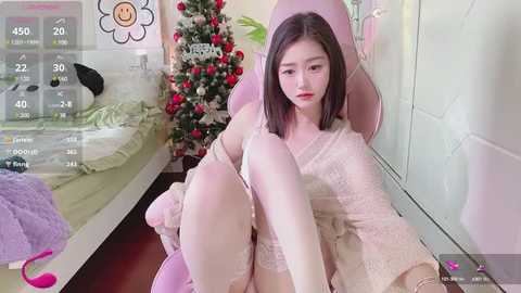 Media: Video of a young East Asian woman with straight black hair, wearing a pink robe, seated on a pink gaming chair. Background features a decorated Christmas tree and a monitor displaying gaming stats.