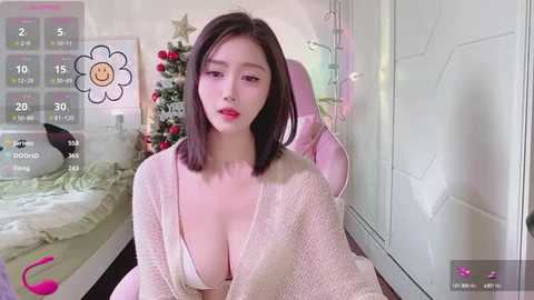 Media: A video of an Asian woman with straight, shoulder-length black hair, fair skin, and large breasts, wearing a white knit cardigan, sitting on a pink gaming chair in a bedroom adorned with a Christmas tree.