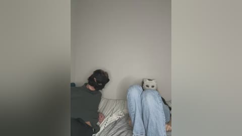 Media: A video of a person in a dark mask and green shirt, seated on a bed with light-colored bedding and blue jeans, with a stuffed owl nearby in a dimly lit room.