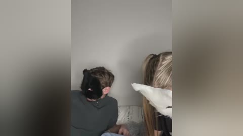 Media: Video of a young man in a Batman mask and gray shirt, sitting on a white couch, facing a blonde woman with a white pillow.