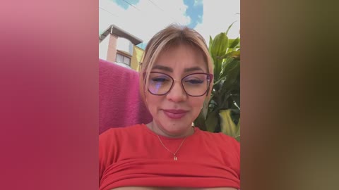 Media: Video of a young Caucasian woman with light skin, blonde hair, wearing glasses and a red shirt, seated outdoors with green foliage and a pink wall in the background.