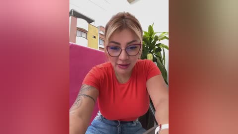 Media: Video of a light-skinned woman with blonde hair in glasses, wearing a red T-shirt and high-waisted jeans, sitting outdoors by a pink wall, with a green plant and multi-colored buildings in the background.