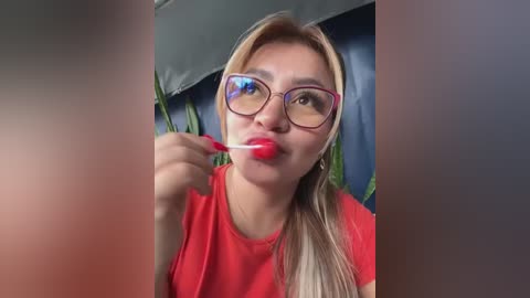 Media: A video of a light-skinned woman with long blonde hair and large purple-framed glasses, wearing a red top, brushing her teeth with red lipstick on her lips. Background shows blurred blue fabric and green plants.