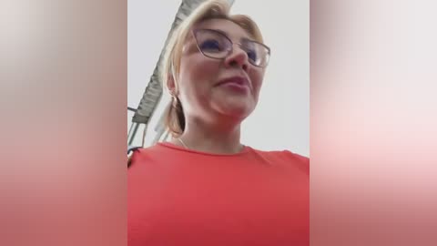 Media: A video of a blonde woman with glasses and a red top, taken from a low angle, with blurred vertical edges.