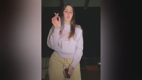 Media: Video of a young woman with long dark hair, wearing a light purple sweater and yellow pants, holding a cigarette, indoors with dim lighting.