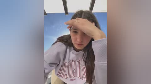 Media: Video of a young woman with long dark hair, wearing a grey sweatshirt with \"Dad\" in red, covering her face with one hand. Background shows a blue sky and white ceiling.