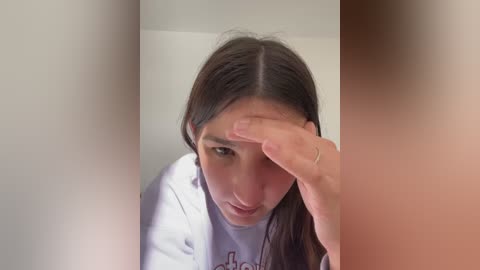 Media: Video of a young woman with long, dark hair, wearing a white shirt, covering her forehead with her right hand, standing in a room with white walls and soft lighting.